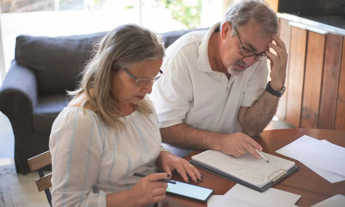 Older couple at a dining table reviewing paperwork on a clipboard, with a smartphone calculator open