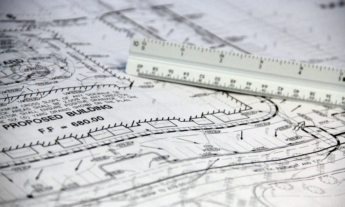An engineer’s scale ruler resting on a printed civil site plan showing lot lines, a proposed building and grading notes